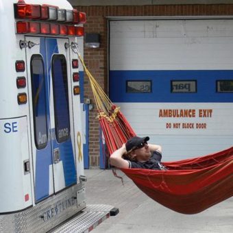 A person sits in a hammock. with their arms behind their head, attached to an ambulance. Ambulance garage behind.