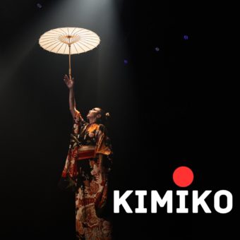 Kaylie Kreatrix stands on a stage reading towards an oiled paper umbrella. In a white font, "Kimiko" with a red circle ontop of the "i" is in the lower right corner.