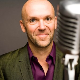 Bremner stands in front of microphone. Grey background.