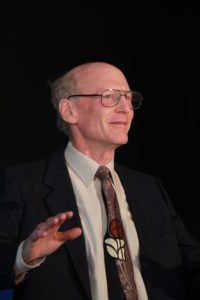 Nicholas Rice stands in a suit and glasses, gesturing with their hand towards the audience. Black background.