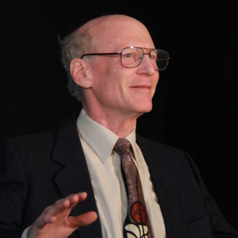 Nicholas Rice stands in a suit and glasses, gesturing with their hand towards the audience. Black background.