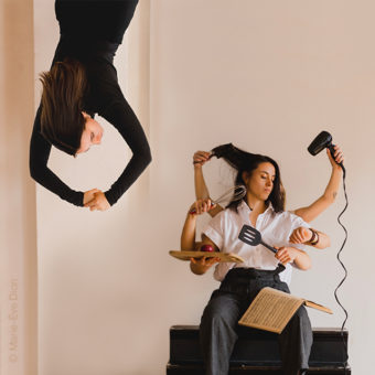 One person appears to be hanging from ceiling. The next person is sitting down with various arms coming from their back holding various objects, such as a blowdryer, a music book on their lap, kitchen utensils and an apple on a cutting board. Cream coloured room.