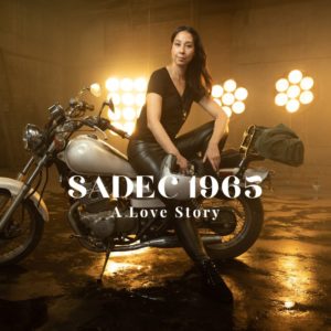 Artist Flora Le sits on a motorcycle. The text reads "Sadec 1965: A Love Story"