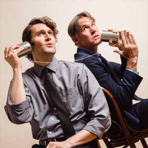 two actors sit side by side holding connected tin cans to their ears.