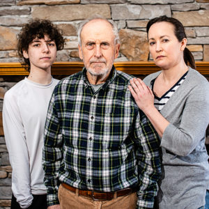 Three members of a family stand next to each other with intense expressions. Bricks and wooden mantel piece behind.
