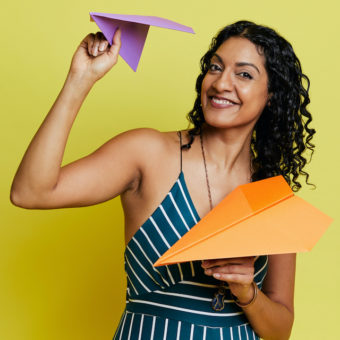 Aliya stands and smiles in front of a yellow background with a purple and orange paper airplane in their hands.